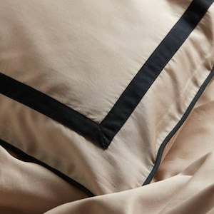 Peut inclure: Un gros plan d'une taie d'oreiller beige avec une bordure noire autour des bords. La taie d'oreiller est faite d'un tissu doux et lisse.
