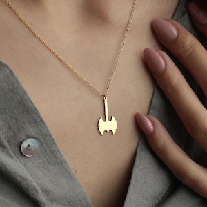 May include: A gold-colored necklace with a small axe pendant hangs on a person's neck. A second, shorter gold chain sits higher on the neck. The person is wearing a gray button-down shirt. The person's hand is visible.