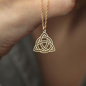 May include: A gold-coloured necklace with a Celtic knot pendant. The pendant has a triangular shape with an interwoven design. The chain is delicate and the necklace is held against a neutral background.