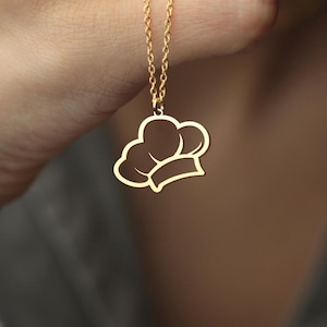 May include: A gold-colored necklace featuring a pendant shaped like a chef's hat. The pendant is a simple outline of the hat design, suspended from a delicate chain. The necklace is held up against a gray background.