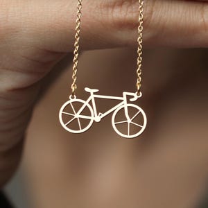 K&ouml;nnte beinhalten: Eine goldfarbene Fahrrad-Anh&auml;nger-Halskette. Der Anh&auml;nger ist ein Ausschnitt eines Fahrrads mit detaillierten R&auml;dern und Rahmen. Die Kette ist eine zarte Goldkette. Die Halskette wird vor einem verschwommenen Hintergrund pr&auml;sentiert.