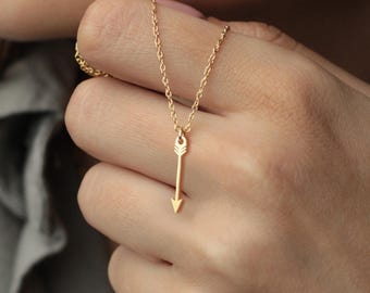 Könnte beinhalten: Eine zarte goldfarbene Pfeil-Anhänger-Halskette. Der Pfeil-Anhänger hängt an einer feinen Kette. Die Halskette wird von einer Hand gehalten. Der Pfeil zeigt nach unten.