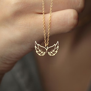 Puede incluir: Un collar dorado con un colgante en forma de alas de &aacute;ngel. Las alas est&aacute;n detalladas con l&iacute;neas delicadas, y la cadena es fina y elegante. El collar se muestra sobre un fondo borroso.