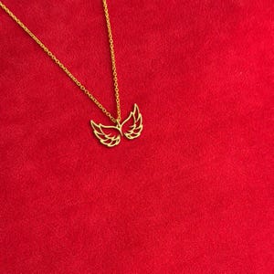 Puede incluir: Un collar dorado con un colgante en forma de alas abiertas. Las alas est&aacute;n detalladas con l&iacute;neas delicadas. La cadena del collar tambi&eacute;n es dorada y descansa sobre un fondo rojo. El colgante es un s&iacute;mbolo de libertad.