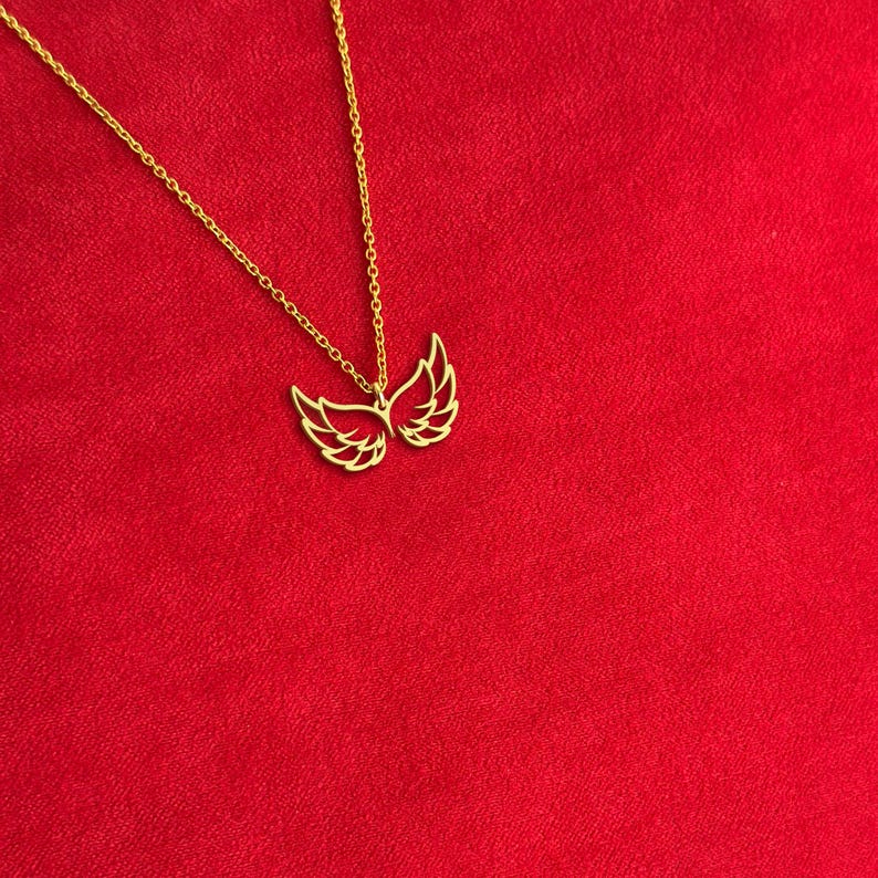 Puede incluir: Un collar dorado con un colgante en forma de alas abiertas. Las alas est&aacute;n detalladas con l&iacute;neas delicadas. La cadena del collar tambi&eacute;n es dorada y descansa sobre un fondo rojo. El colgante es un s&iacute;mbolo de libertad.