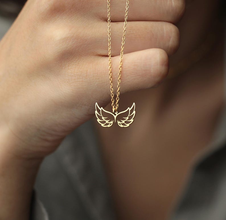 Puede incluir: Un collar dorado con un colgante en forma de alas de &aacute;ngel. Las alas est&aacute;n detalladas con l&iacute;neas delicadas, y la cadena es fina y elegante. El collar se muestra sobre un fondo borroso.