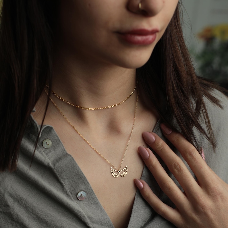 Puede incluir: Collar dorado con un delicado colgante de alas. Se lleva con una cadena m&aacute;s fina encima. La persona de la imagen lleva una camisa gris abotonada. Joyer&iacute;a.