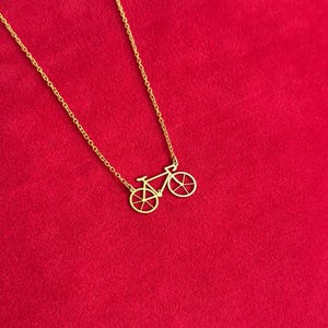 K&ouml;nnte beinhalten: Eine goldfarbene Fahrrad-Anh&auml;nger-Halskette. Der Anh&auml;nger ist ein detaillierter Ausschnitt eines Fahrrads. Die Halskette und der Anh&auml;nger sind goldfarben. Der Hintergrund ist einfarbig rot.