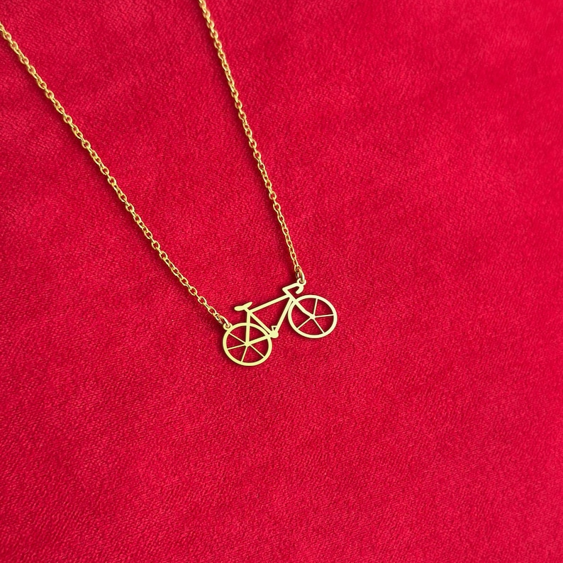 K&ouml;nnte beinhalten: Eine goldfarbene Fahrrad-Anh&auml;nger-Halskette. Der Anh&auml;nger ist ein detaillierter Ausschnitt eines Fahrrads. Die Halskette und der Anh&auml;nger sind goldfarben. Der Hintergrund ist einfarbig rot.