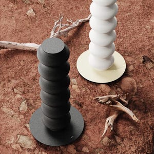 Puede incluir: Dos esculturas decorativas modernas, una negra y otra blanca, ambas con un diseño apilado y redondeado. La escultura negra se asienta sobre una base gris, mientras que la blanca descansa sobre una base dorada. El fondo es una superficie texturizada de color marrón rojizo.