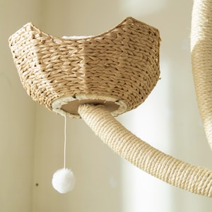 May include: A cat tree with a woven basket-style bed and a rope-wrapped post. A white pom-pom toy hangs from the bed. The cat tree is made of natural materials and is designed for indoor use.
