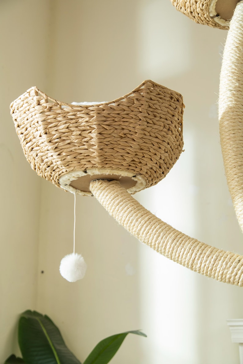May include: A cat tree with a woven basket-style bed and a rope-wrapped post. A white pom-pom toy hangs from the bed. The cat tree is made of natural materials and is designed for indoor use.