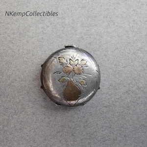 May include: A silver-toned, round antique compact with a floral design. The compact features an engraved vase with gold-toned flowers. The top has a small, textured knob. The text "NKempCollectibles" is in the upper left corner.