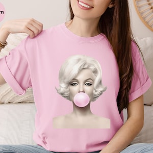 May include: A light pink t-shirt featuring a black and white image of Marilyn Monroe blowing a bubblegum bubble. The word "Blossom" is in a pink circle in the upper left corner.