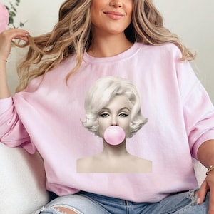 Puede incluir: Sudadera rosa claro con una imagen en blanco y negro de Marilyn Monroe soplando una burbuja rosa. La sudadera es de color rosa claro liso. Las palabras "Light Pink" están en un círculo rosa en la esquina superior izquierda.