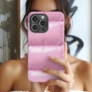 May include: A pink puffer jacket style phone case. The case has a glossy finish and a quilted design. The phone case has a black camera module with three lenses and a flash. The case is held in front of a person.