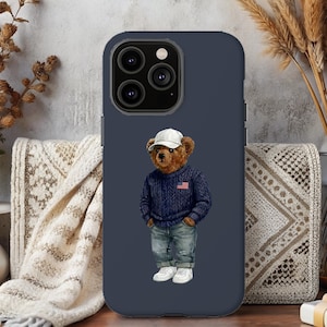 May include: Navy blue phone case featuring a cartoon bear wearing a blue sweater with an American flag, jeans, a white cap, and sneakers. The case has a dark blue border and a camera cutout.