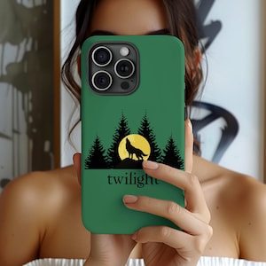 May include: Green phone case featuring a silhouette of a howling wolf against a yellow moon, with pine trees and the word "twilight" below. The case is held up in front of a person's face.