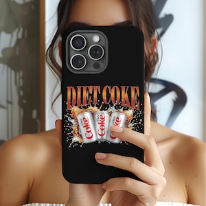 May include: Black phone case featuring the Diet Coke logo in orange and white. The design includes three Diet Coke cans with a splash effect. The case is held up in front of a person.