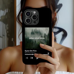 May include: Black phone case featuring a forest image with the text "HOA HOA HOA." The phone displays a music player interface with the song "Eyes On Fire" by Blue foundation. The case has a sleek, modern design.