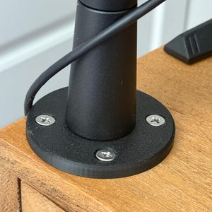 May include: Close-up of a black monitor stand base with three silver screws securing it to a wooden surface.