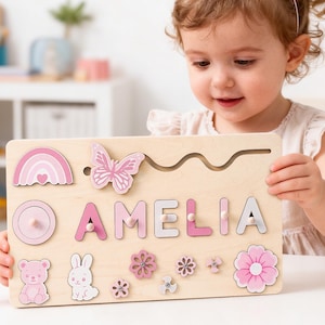 Puede incluir: Un rompecabezas de madera con el nombre "AMELIA" en letras rosas y blancas. El rompecabezas incluye un arcoíris, una mariposa, flores, un oso y un conejo. El niño lleva un gorro de cumpleaños.