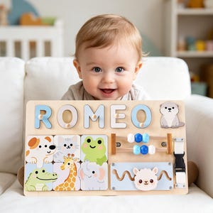 May include: A wooden activity board featuring the name ROMEO in light blue letters, a cartoon otter, and six square tiles with illustrations of a dog, cat, frog, crocodile, giraffe, and elephant. It includes a blue bead abacus and a sliding llama.