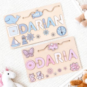 K&ouml;nnte beinhalten: Zwei Holz-Namenspuzzles, eines mit der Aufschrift "DARIAN" in Blau und das andere mit "DARIA" in Rosa. Jedes Puzzle hat dekorative Formen wie einen Wal, ein Segelboot und Blumen. Die Puzzles liegen auf einer wei&szlig;en Oberfl&auml;che.