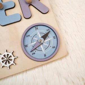 K&ouml;nnte beinhalten: Holzpuzzle mit einer hellblauen Windrose, einem wei&szlig;en Steuerrad und hellblauen und lila Buchstaben. Der Kompass hat eine schwarze Nadel und ist vor einem hellblauen Hintergrund angeordnet. Die Buchstaben sind in einer verspielten Schriftart.