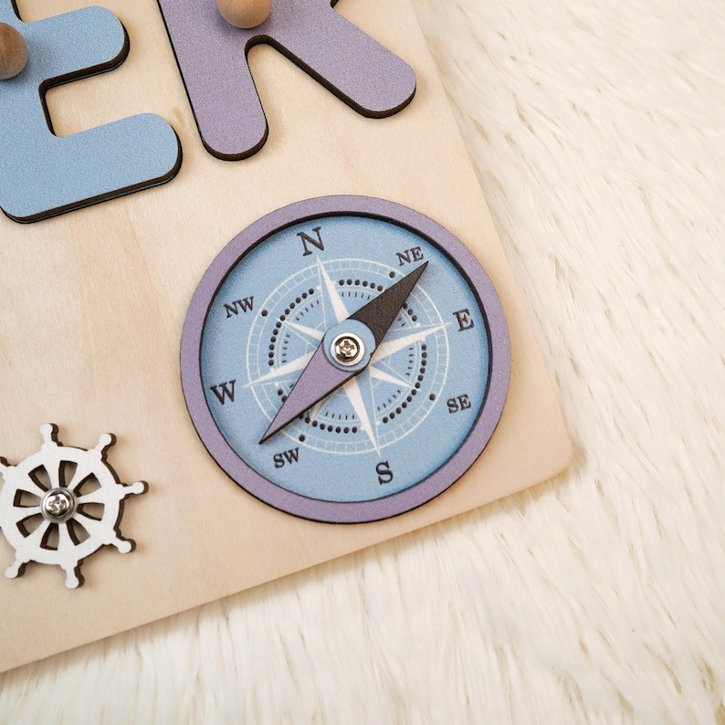 K&ouml;nnte beinhalten: Holzpuzzle mit einer hellblauen Windrose, einem wei&szlig;en Steuerrad und hellblauen und lila Buchstaben. Der Kompass hat eine schwarze Nadel und ist vor einem hellblauen Hintergrund angeordnet. Die Buchstaben sind in einer verspielten Schriftart.