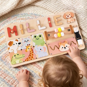 May include: A wooden activity board featuring the name MILLIE in colorful letters, a cartoon dog, a bead maze with yellow and white beads, a black buckle strap, and a sliding track with a squirrel piece. Below are square tiles of animals.