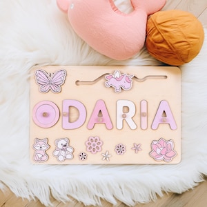 K&ouml;nnte beinhalten: Holz-Namenspuzzle mit dem Namen "ODARIA" in rosa und wei&szlig;en Buchstaben. Das Puzzle enth&auml;lt einen Schmetterling, ein Einhorn, einen Fuchs, einen Teddyb&auml;ren und Blumen. Das Puzzle befindet sich auf einer hellen Holzbasis.