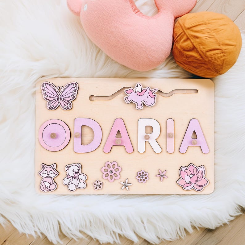 K&ouml;nnte beinhalten: Holz-Namenspuzzle mit dem Namen "ODARIA" in rosa und wei&szlig;en Buchstaben. Das Puzzle enth&auml;lt einen Schmetterling, ein Einhorn, einen Fuchs, einen Teddyb&auml;ren und Blumen. Das Puzzle befindet sich auf einer hellen Holzbasis.