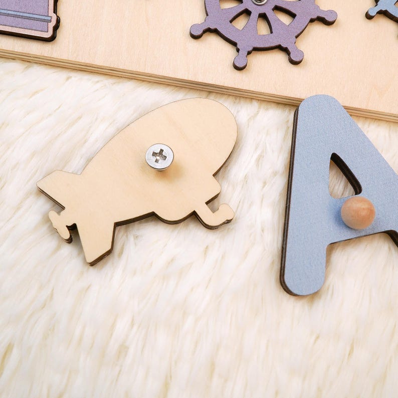 K&ouml;nnte beinhalten: Holzpuzzle mit verschiedenen Formen und Buchstaben. Enth&auml;lt eine hellbraune Rakete, ein lila Steuerrad und den hellblauen Buchstaben "A". Die Puzzleteile haben Holzkn&ouml;pfe. Der Hintergrund ist eine flauschige, wei&szlig;e Oberfl&auml;che.