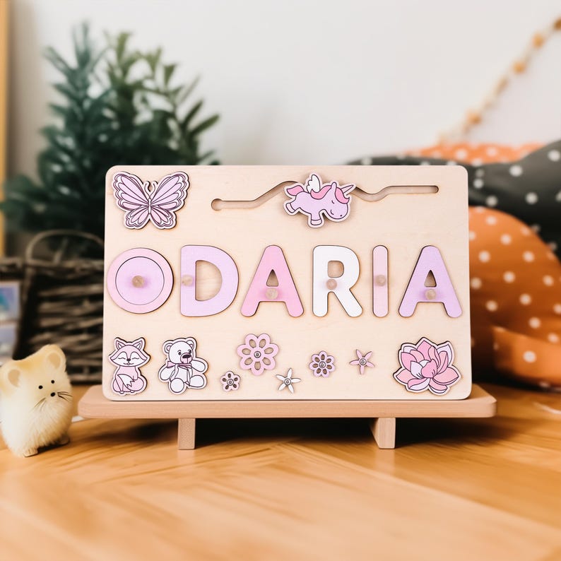K&ouml;nnte beinhalten: Holz-Namenspuzzle mit dem Namen "ODARIA" in rosa und wei&szlig;en Buchstaben. Das Puzzle zeigt einen Schmetterling, ein Einhorn, einen Fuchs, einen Teddyb&auml;ren, Blumen und eine Lotusblume. Das Puzzle steht auf einem kleinen Holzst&auml;nder.