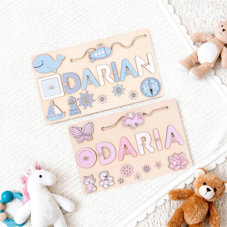 K&ouml;nnte beinhalten: Zwei Holz-Namenspuzzles, eines mit der Aufschrift "DARIAN" in Blau und das andere mit "DARIA" in Rosa. Jedes Puzzle hat dekorative Formen wie einen Wal, ein Segelboot und Blumen. Die Puzzles liegen auf einer wei&szlig;en Oberfl&auml;che.