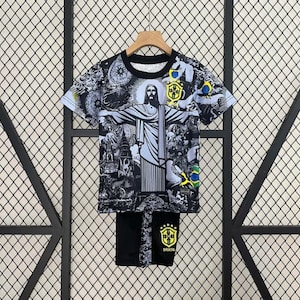 Puede incluir: Conjunto de camiseta y pantal&oacute;n corto con gr&aacute;ficos en blanco y negro con la estatua del Cristo Redentor y otras im&aacute;genes brasile&ntilde;as. La camiseta tiene mangas cortas y cuello redondo. El pantal&oacute;n corto lleva la palabra "Brasil" y un escudo. El conjunto se muestra en una percha.