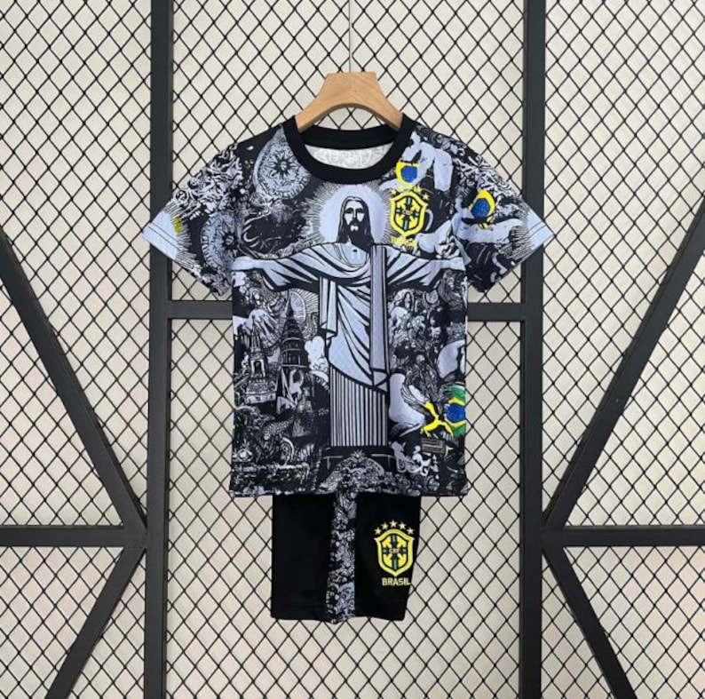 Puede incluir: Conjunto de camiseta y pantal&oacute;n corto con gr&aacute;ficos en blanco y negro con la estatua del Cristo Redentor y otras im&aacute;genes brasile&ntilde;as. La camiseta tiene mangas cortas y cuello redondo. El pantal&oacute;n corto lleva la palabra "Brasil" y un escudo. El conjunto se muestra en una percha.