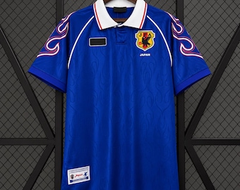 Puede incluir: Un polo azul de manga corta con cuello blanco y ribete rojo, patrones de llamas en las mangas y el escudo de JFA Japan en el pecho. La tela tiene una textura de llamas tono sobre tono y una etiqueta rectangular visible en la parte inferior.
