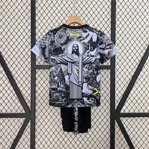 Puede incluir: Conjunto de camiseta y pantal&oacute;n corto con gr&aacute;ficos en blanco y negro con una ilustraci&oacute;n detallada de Cristo Redentor con los brazos extendidos. La camiseta tiene cuello redondo y mangas cortas. El pantal&oacute;n corto es negro. La palabra "BRASIL" es visible en la camiseta.