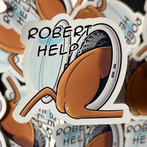 Könnte beinhalten: Ein Aufkleber mit einer Cartoon-Illustration eines braunen Objekts mit einem grauen und weißen Plattenspieler-Design. Der Aufkleber trägt den Text "ROBERT HELP!" in Schwarz mit weißem Rand. Der Hintergrund ist mit ähnlichen Aufklebern gefüllt.
