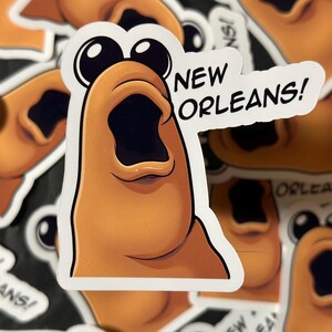 Könnte beinhalten: Ein Aufkleber mit einer cartoonartigen, überrascht aussehenden Kreatur in Orange- und Brauntönen, mit großen schwarzen Augen und offenem Mund. Der Aufkleber hat die Worte "NEW ORLEANS!" in einer Sprechblase.