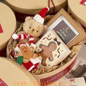 Könnte beinhalten: Eine weihnachtliche Geschenkbox mit einem Teddybär mit Weihnachtsmütze und Schal, einem gerahmten Bild eines Weihnachtsbaums und Lebkuchen-Artikeln. Die Box ist rund mit einem rot-weißen Streifen.
