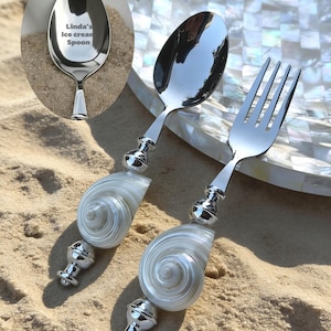 May include: A stainless steel ice cream spoon and fork set with seashell handles. The spoon has the text "Linda's Ice cream Spoon" engraved on the bowl. The handles are white and silver, and the set is displayed on sand.