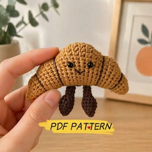 May include: A crocheted croissant with a smiling face and dark brown legs. The pastry is light brown with darker brown stripes. The text "PDF PATTERN" is in a yellow banner below the croissant.