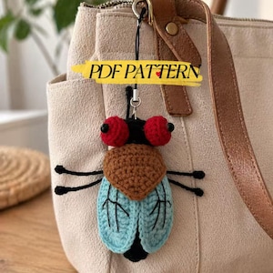 May include: A handmade crochet fly keychain with red eyes, a brown head, and blue wings. The fly is attached to a beige handbag. The text "PDF PATTERN" is on a yellow banner.