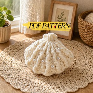 May include: Cream-colored, crocheted seashell with pearl accents. The shell is placed on a beige, crocheted doily. The image also includes a small potted plant, a framed picture, and a woven basket.