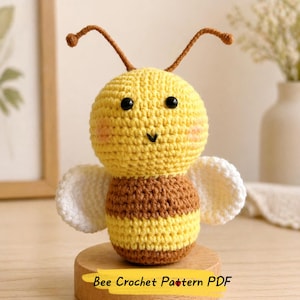 May include: A crocheted bee toy, featuring a yellow head, a brown and yellow striped body, and white wings. The bee has black eyes, pink cheeks, and brown antennae. The text "Bee Crochet Pattern PDF" is visible.