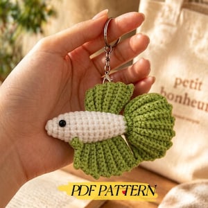 May include: A crochet keychain shaped like a betta fish with a white body, a black bead eye, and textured green fins. The fish is attached to a silver-colored metal keyring and chain. Below the fish, the text PDF PATTERN is printed in black.