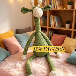 May include: A handmade green and white crochet giraffe with long legs and a long neck. The giraffe has white accents on its head and feet. The text "PDF PATTERN" is across the giraffe's body. The giraffe is sitting on a pink fluffy rug.